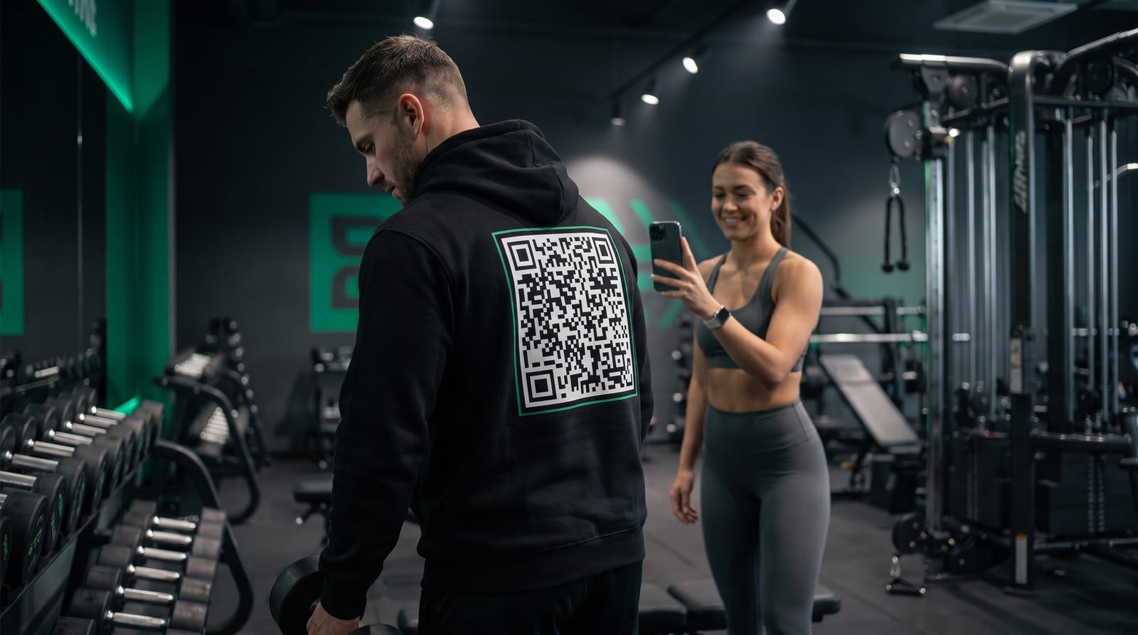 Person in a gym wearing a hoodie with a QR code on the back while someone else scans it
